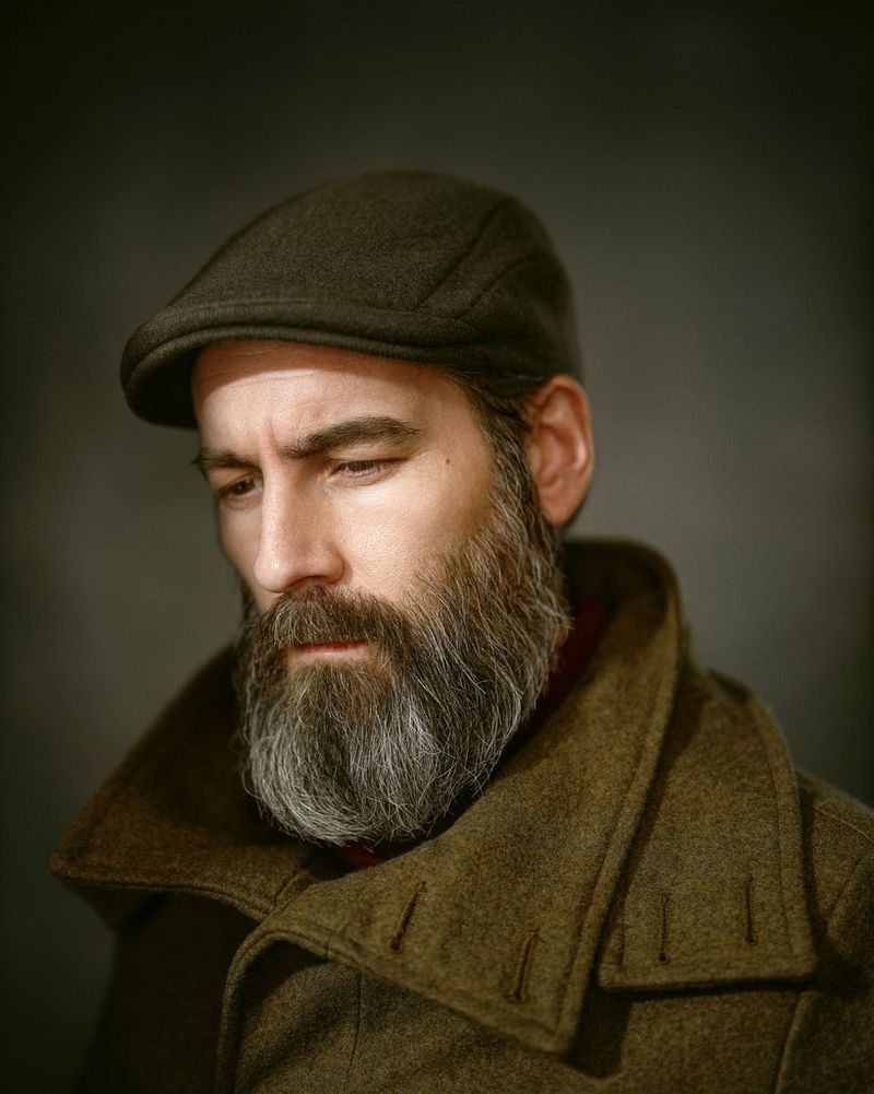 Un uomo con la barba guarda verso l'angolo in basso della fotocamera, con gli occhi quasi chiusi. Indossa un cappotto marrone in lana con un ampio colletto e un basco marrone scuro.