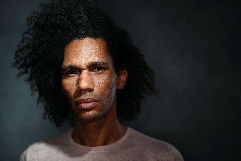 Un uomo con folti capelli neri ricci e una maglia bianca fissa intensamente l'obiettivo contro uno sfondo grigio scuro.