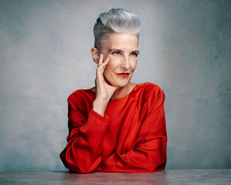 Una donna con capelli argentati e una maglia in seta rossa posa il viso sulla mano mentre si appoggia elegantemente a un tavolo. Sta guardando in lontananza.