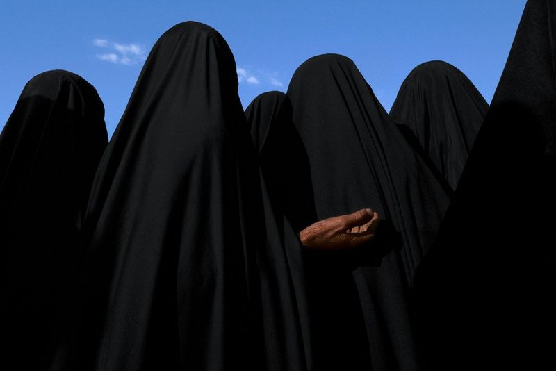 A group of five people stand against the blue sky, dressed in black burqas. The person closest to the camera raises their right hand. This photograph was captured by James Nachtwey in Karbala, Iraq in 2003.
