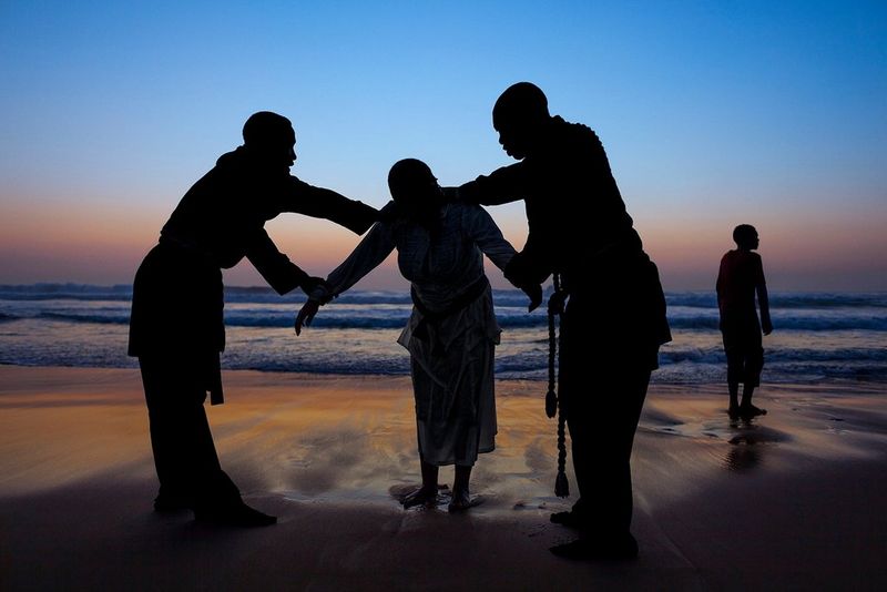 Four people stand on a beach in South Africa. Three of them stand together, as two of the people hold the person in the middle. There is a fourth person in the background. This photograph was captured by James Nachtwey. 