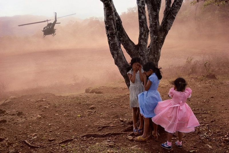 Three little girls dressed in white, blue and pink clothes stand by a tree, covering their eyes to shield themselves from the dust displaced by a helicopter flying in the distance. This photograph was captured by James Nachtwey in San Luis de la Reina, El Salvador in 1984.