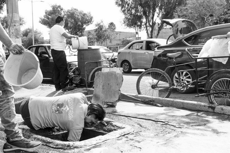 Черно-белое фото, созданное Жеромом Сессини на камеру Canon EOS R5, демонстрирует человека, который лежит на земле и сунул руку в яму на улице. Другие люди поблизости несут ведра, а по дороге едут машины.