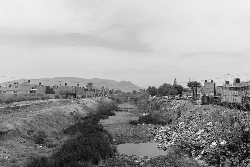 Черно-белое фото, созданное Жеромом Сессини на камеру Canon EOS R5, демонстрирует пересохшую реку в Экатепеке, Мексика, и здания по обоим берегам.