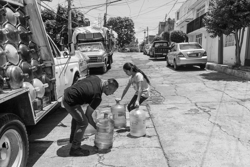 Черно-белое фото, созданное Жеромом Сессини на камеру Canon EOS R5, демонстрирует продавца воды и девочку с большими бутылями воды на улице, а также припаркованный рядом грузовик с такими же бутылями.