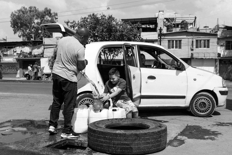 Черно-белое фото, созданное Жеромом Сессини на камеру Canon EOS R5, демонстрирует мужчину, наливающего воду из ведра в бутылку, которую держит в руках ребенок, сидящий на улице возле белой машины.