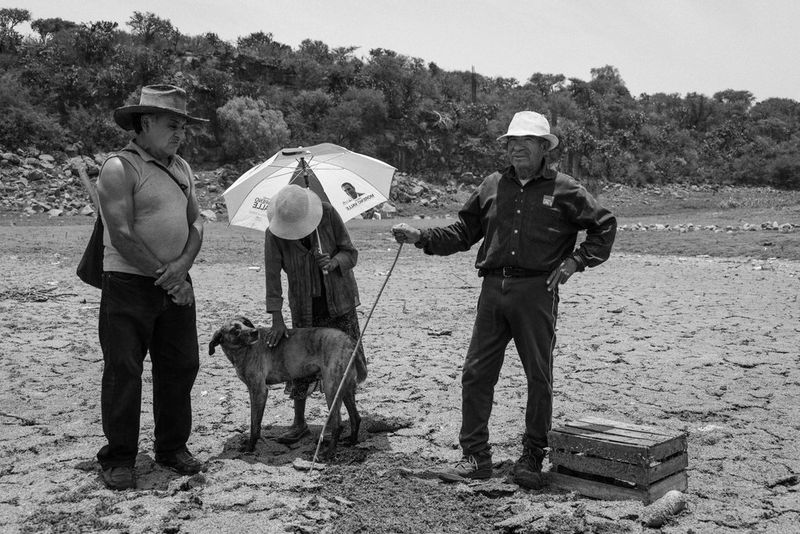 Черно-белое фото, созданное Жеромом Сессини на камеру Canon EOS R5, демонстрирует двух мужчин и женщину, стоящих среди пода высохшего озера. Женщина держит в одной руке зонт, а второй тянется к собаке, чтобы погладить ее.