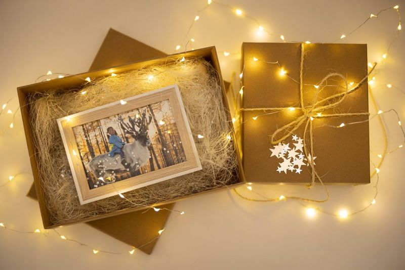 A framed picture of a girl on a reindeer sits in a brown gift box, scattered with metallic confetti and surrounded by festive lights.