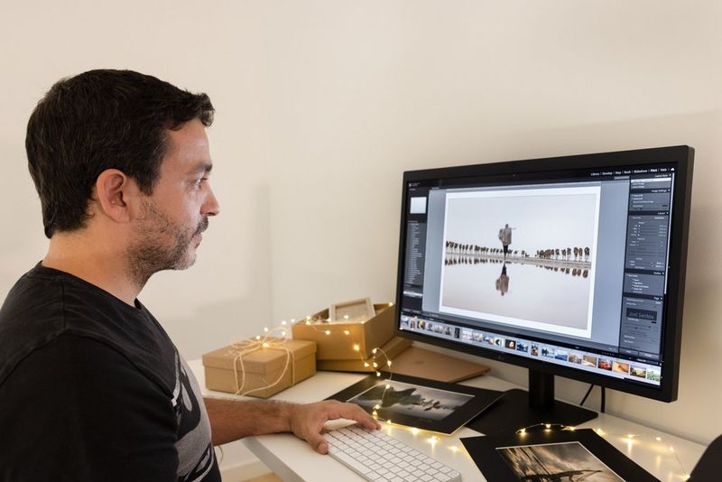 A man sits at a desk editing a photograph on a computer monitor. Gift boxes, festive lights and mounted prints are on the desk beside him.