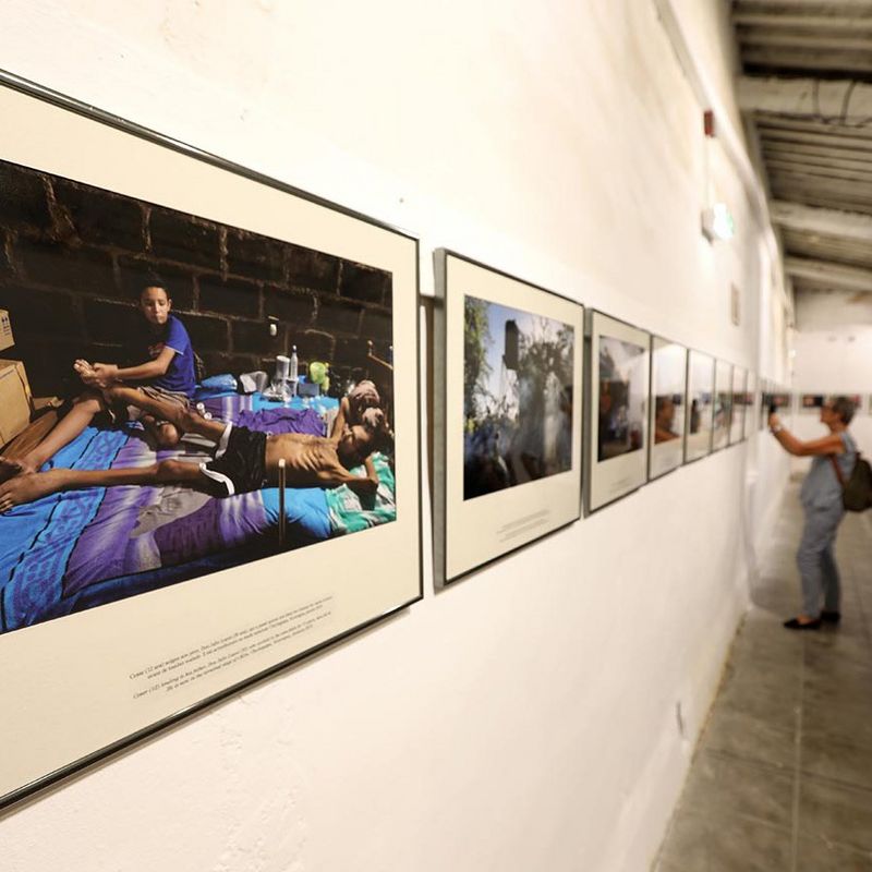 A close-up of a wall of framed photos, with a viewer taking a photograph of a picture in the distance.