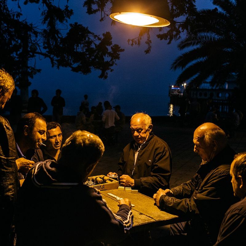 A group of Abkhaz men sit around a table near the sea playing dominoes under a lamp.