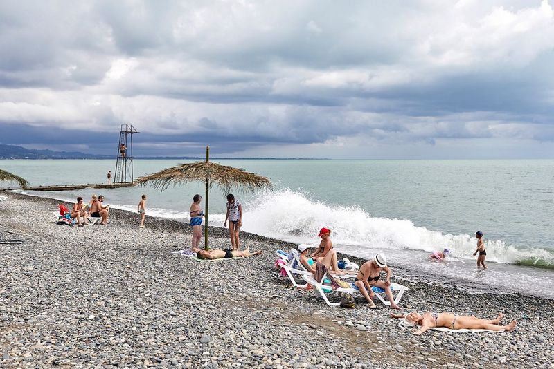 Beneath a cloudy sky, tourists sit on a pebble beach in Abkhazia, while children play in the waves.