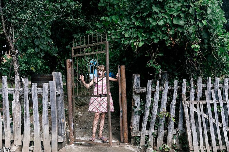 A young village girl in a short pink dress stands behind a rusty metal gate. She is fascinated by something in the distance.