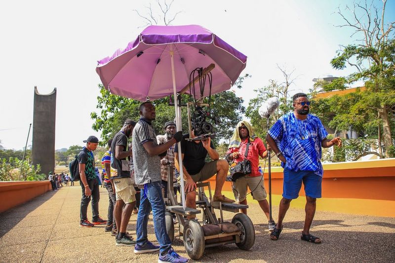 Director Kunle Afolayan and his crew filming outdoors in bright sunshine. A large purple umbrella is being used to shade the camera.