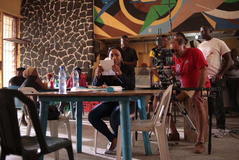 Actor Temi Otedola sits at a canteen table reading a flyer, with a camera crew filming her from behind.