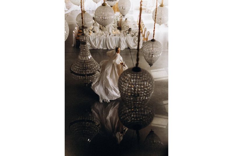 A bride looks up at the camera through ornate pendant lamps hanging in a large room. Environmental portrait taken with a Canon RF 50mm F1.4L VCM lens.