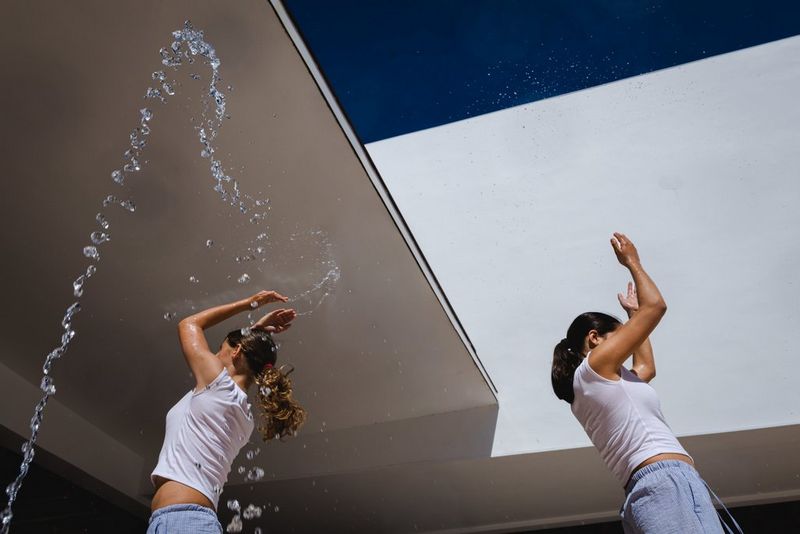 Two young women raise their arms in the air in front of graphic white architecture and a blue sky, with a fountain of water droplets to one side. Action photo taken with a Canon RF 20-50mm F4L IS USM PZ lens.
