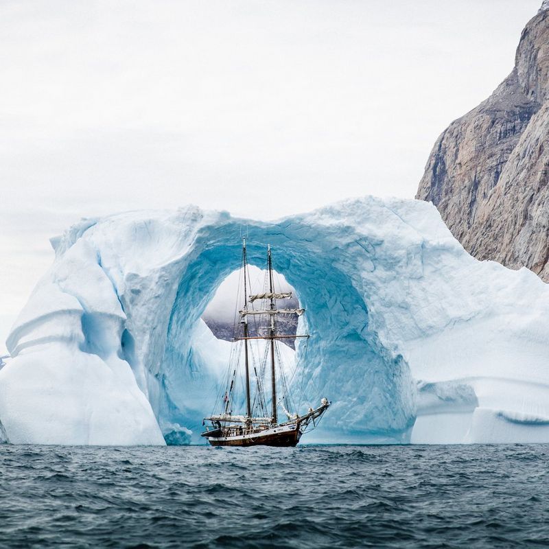 A large sail-powered ship passing through an archway in an iceberg on a rocky coastline. 
