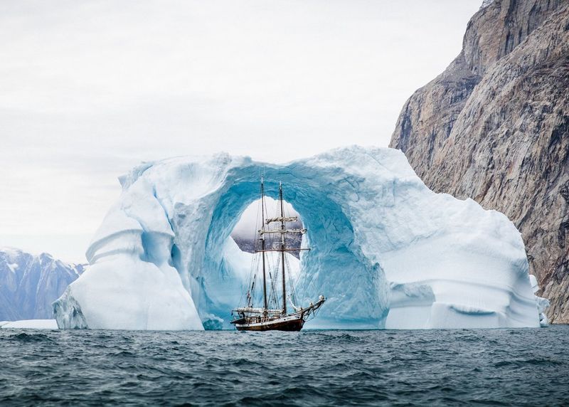Большое парусное судно проходит через арку айсберга на скалистом побережье.