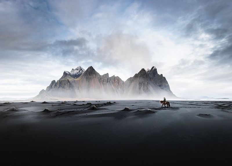 Пейзажное изображение песчано-скалистого образования на черном пляже, по которому два человека едут на лошади.