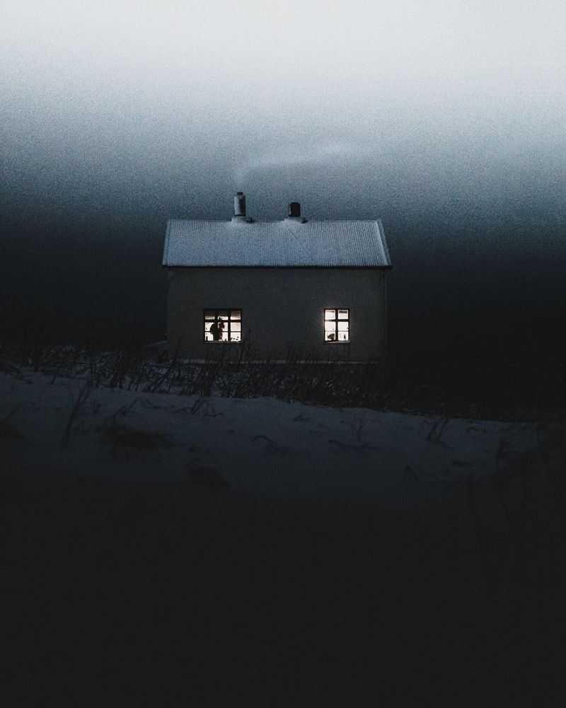 Запечатленный во время сумерек небольшой коттедж со светом в окнах и дымом из трубы стоит среди заснеженного пейзажа.