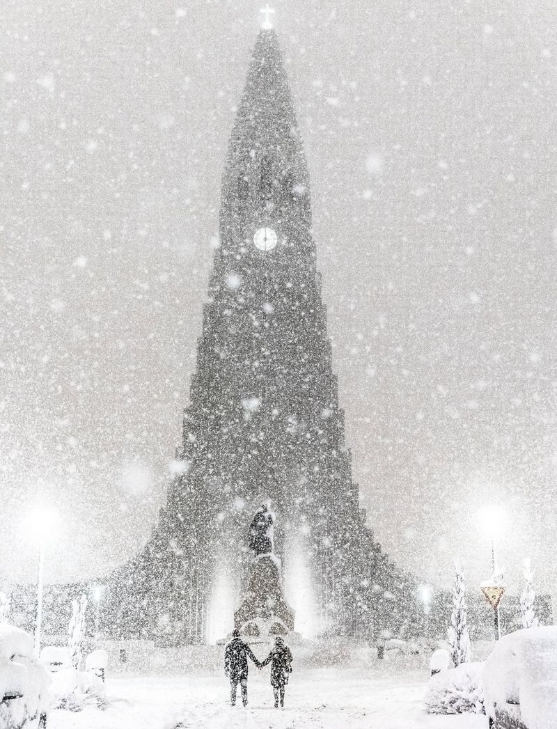 Два человека идут к знаменитой церкви Хадльгримскиркья в сильную метель, держась за руки.