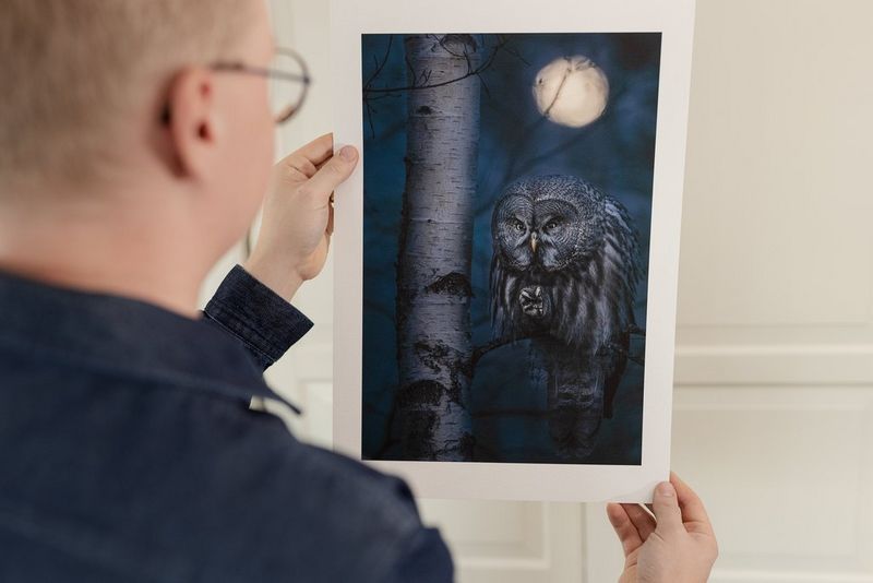 A man holds up a large print of an owl on a branch with the moon overhead.