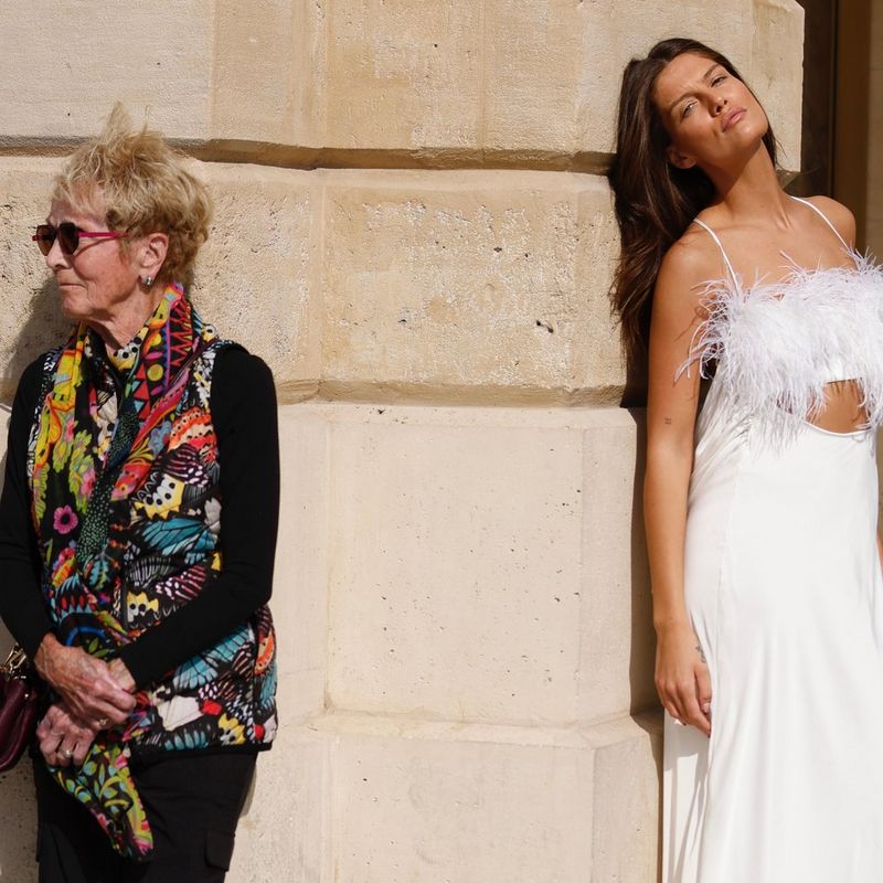 Two women stand against a wall either side of a corner. One is dressed in black everyday clothes with a brightly coloured scarf and gilet, and the other is wearing a long white evening dress. Taken on a Canon EOS R5 by fashion photographer Leo Faria.