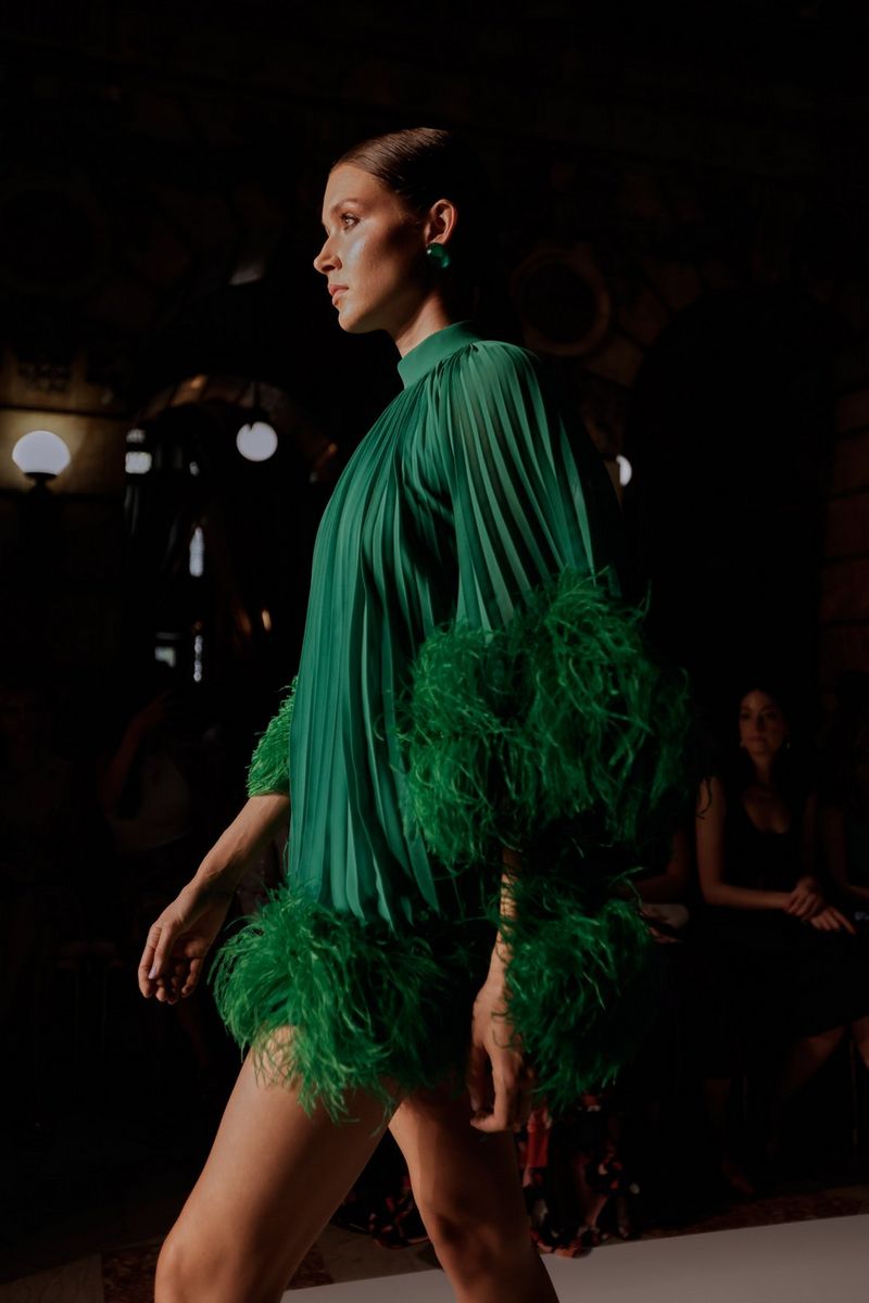 A model walking the runway in a dark green pleated dress with a feather trim. Taken by fashion photographer Leo Faria using the Canon EOS R5. 