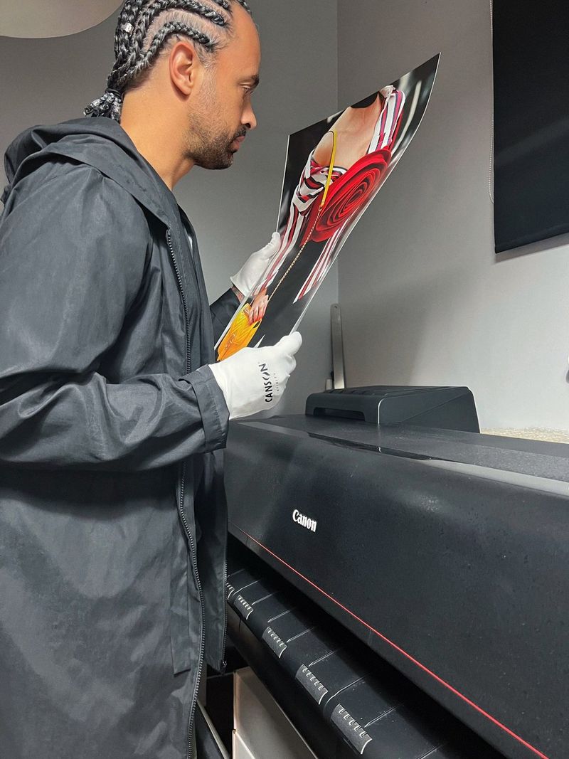 Fashion photographer Leo Faria inspects a print of one of his photographs while wearing white gloves, standing next to a Canon large-format printer. 