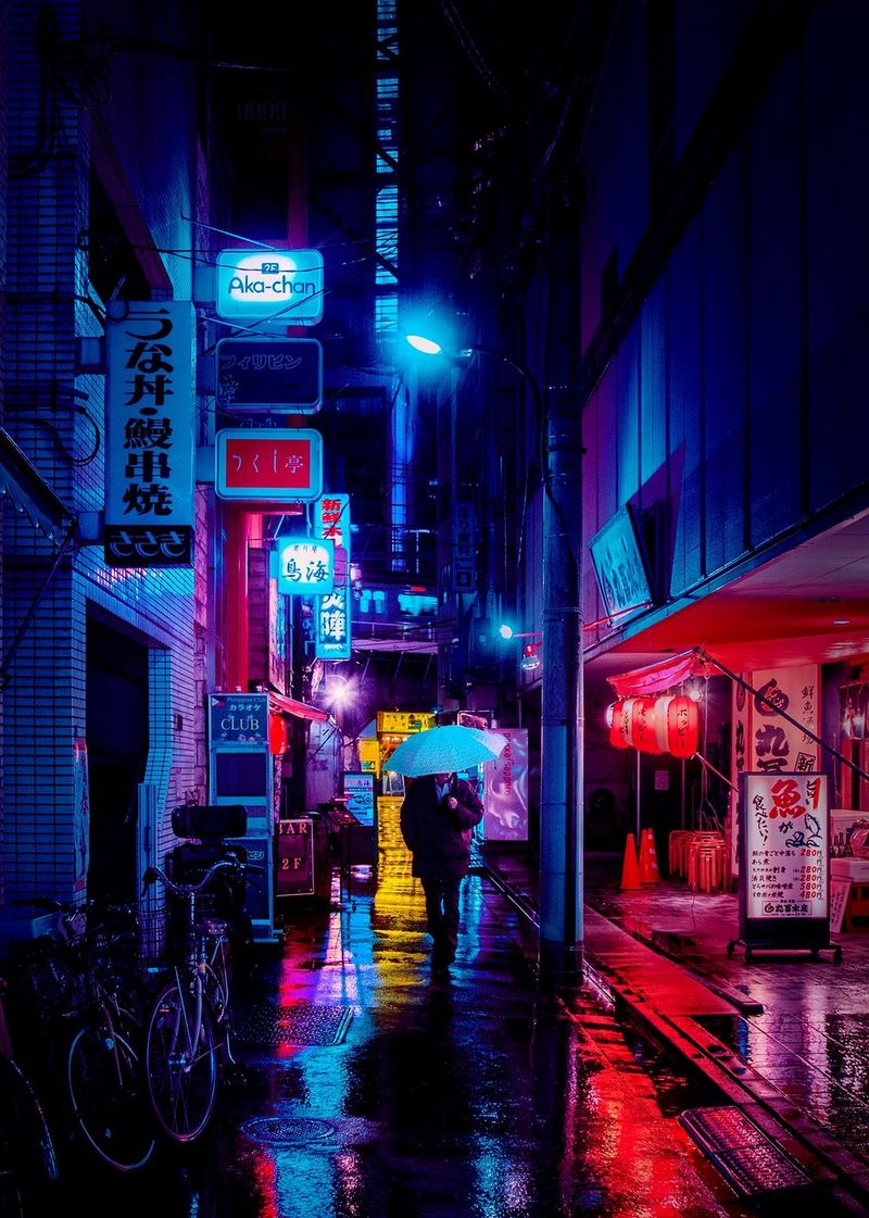 A man holding an open umbrella walks through a neon-lit Tokyo alleyway in the rain at night.