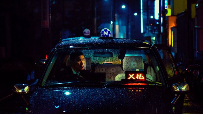A Tokyo taxi driver waits outside a hotel. In the background is a darkened street illuminated by neon signs. 