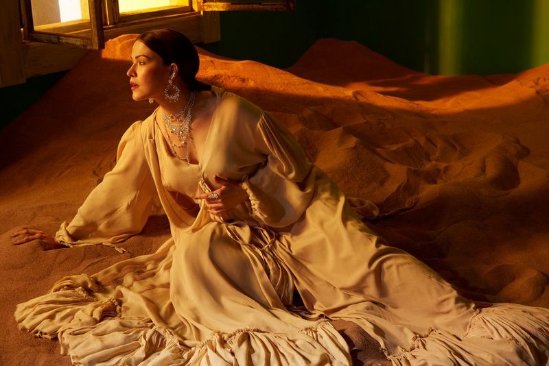 Женщина в белом платье сидит на песке на съемочной площадке, а через окно в комнату, где она находится, проникает свет.