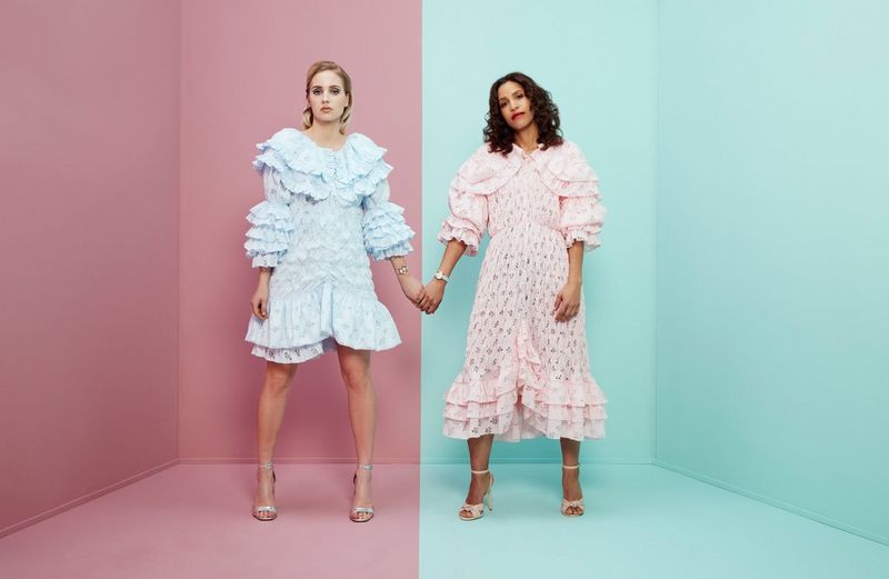 Женщина в синем платье и еще одна женщина в розовом платье стоят в комнате, держась за руки. Комната также разделена на половины — розовую и синюю.