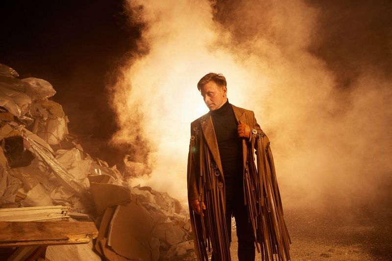 Мужчина в куртке песочного оттенка с длинной бахромой смотрит вниз на кучу мусора, а у него за спиной валит дым.