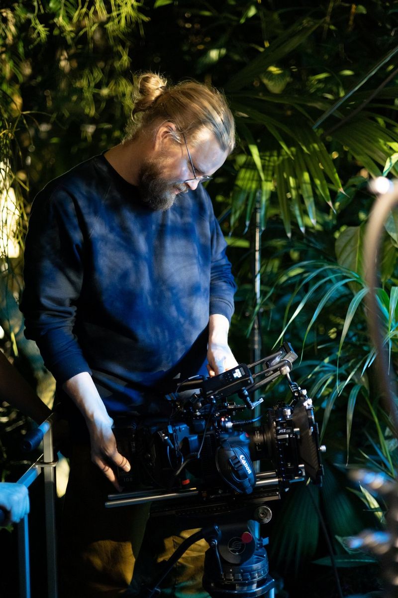 Мужчина в очках держит в руках кинокамеру Canon в окружении зеленых растений.