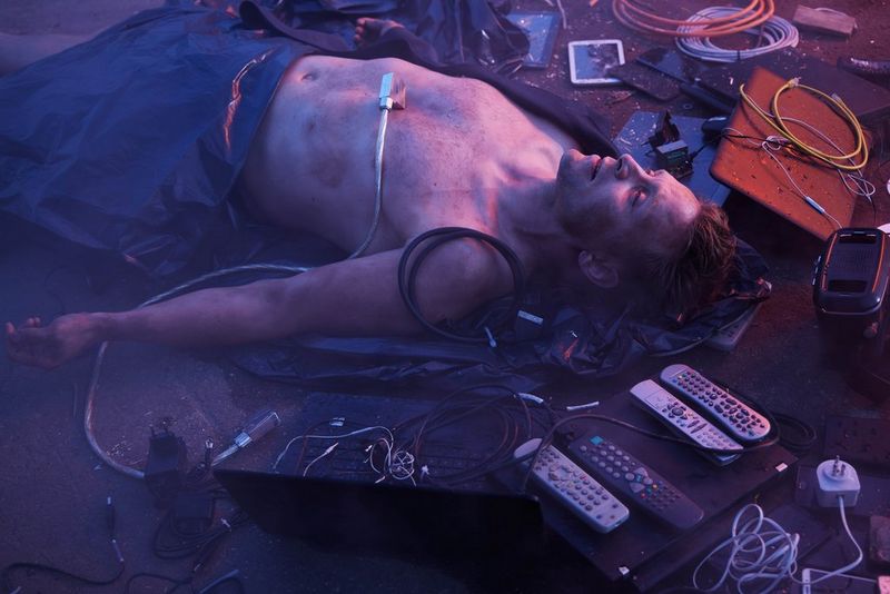 Мужчина без одежды лежит на земле в окружении пультов от телевизора, проводов и других электронных устройств, а нижняя половина его тела покрыта тканью.