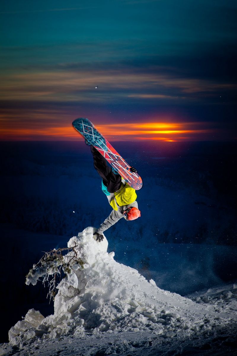 Сноубордист летит вниз головой, а его рука опирается на покрытое снегом дерево, в то время как оранжевое солнце контрастирует за его спиной с бирюзовым небом.