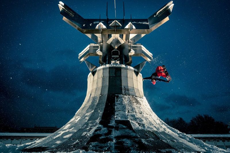 Лаконичная серая скульптура из бетона и металла на фоне звездного ночного неба; сбоку от нее сноубордист выполняет прыжок и летит головой вниз, одной рукой касаясь бетона.