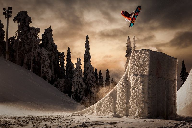 Сноубордист прыгает с покрытой снегом рампы на фоне высоких заснеженных деревьев.