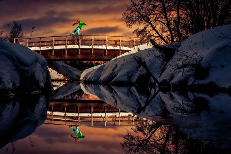 Сноубордист скользит по деревянным перилам моста между двух заснеженных берегов — мост идеально отражается от поверхности воды.