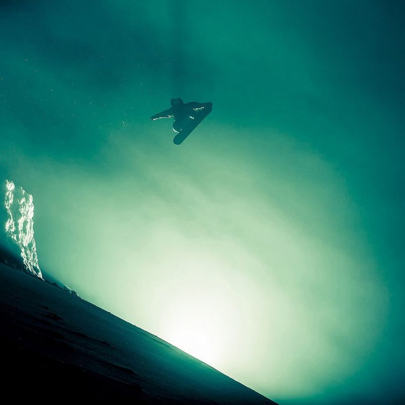 A snowboarder high in the air in foggy conditions, eerily lit from behind the crest of a hill.
