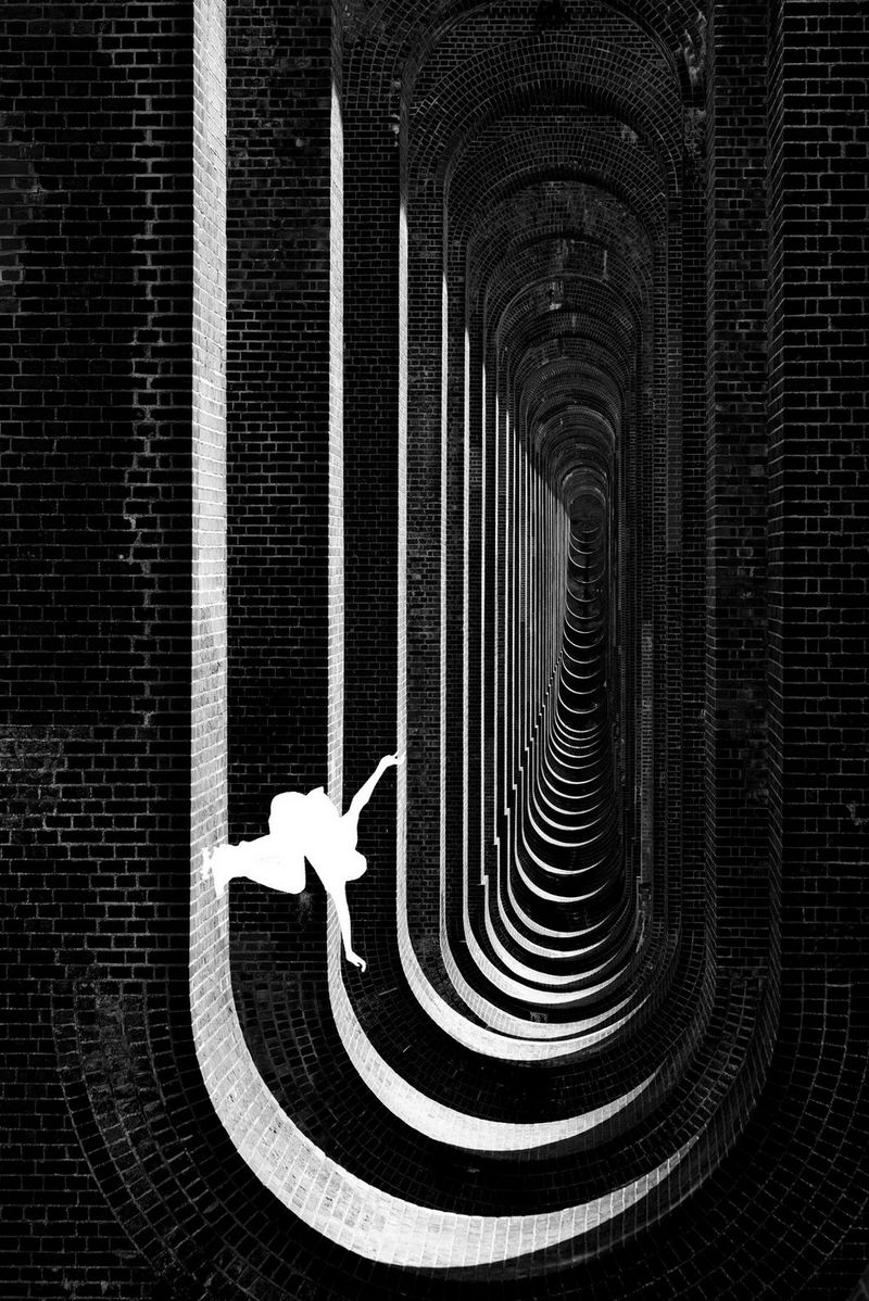 A black and white image shows underneath the viaduct, with a white silhouette figure skateboarding down one side of a wall.