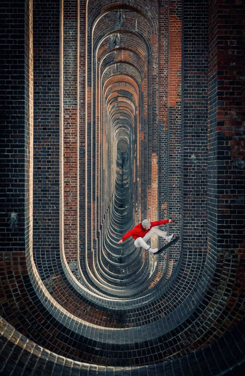 A skateboarder performing a fakie kickflip inside the curved brickwork structure of a viaduct, framed so that the brickwork repeats recursively into the distance.