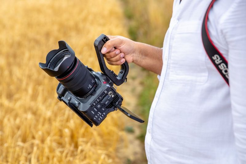 A man in a white shirt standing at the edge of a field holding a Canon EOS C70 by a mounted handle.
