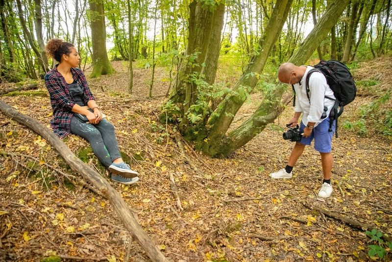 Filmmaker Simeon Quarrie filming British artist, Bryony Benge-Abbott as she sits on a tree stump in the woods.