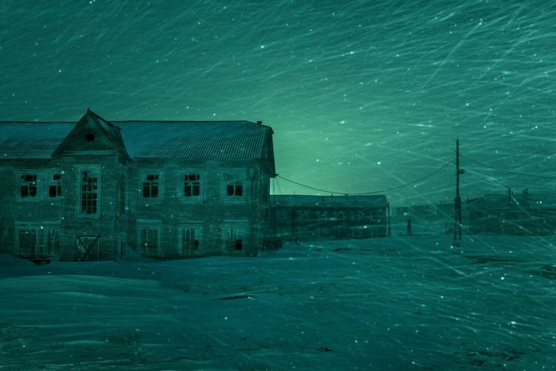 A building in a snowy, Arctic environment lit up in green by the Aurora Borealis. Snowflakes in motion can be seen in the air in the foreground.