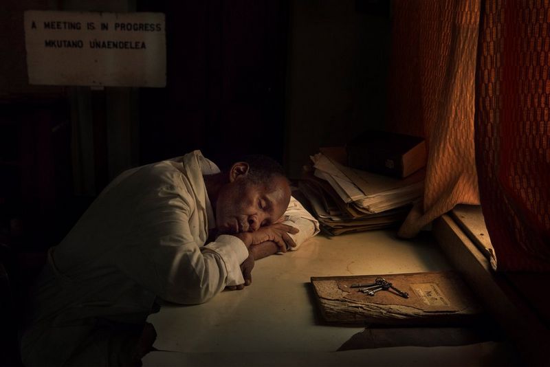 A man in a dark room resting his head on a desk beside a window with orange curtains.