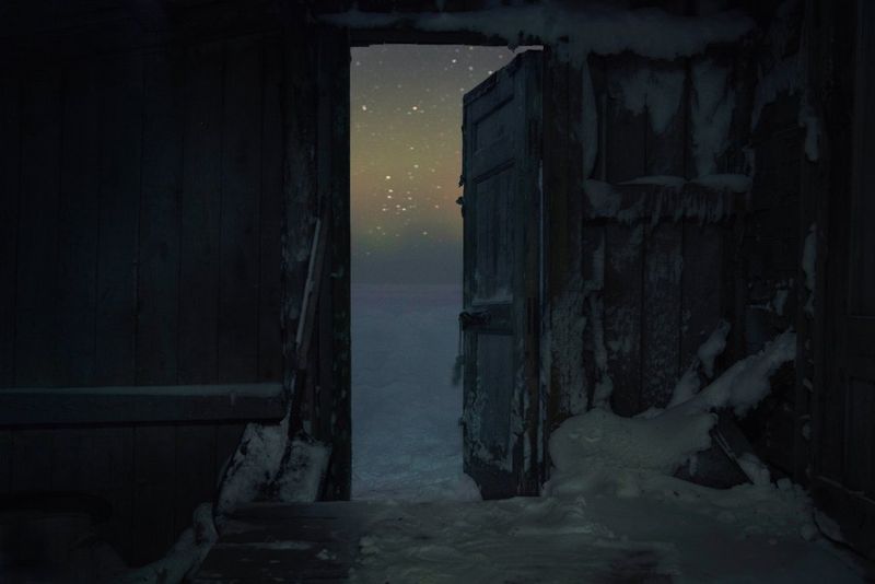 A shot taken in a dark, wooden shed, full of snow. A view of snow and evening sky can be seen through the open door.