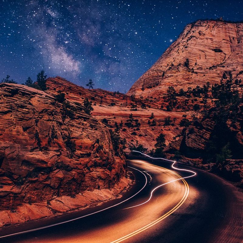 Rocky outcrops in Zion National park, Utah, USA, pictured at night. Blurred lights run along the road and the Milky Way can be seen in the sky above. 